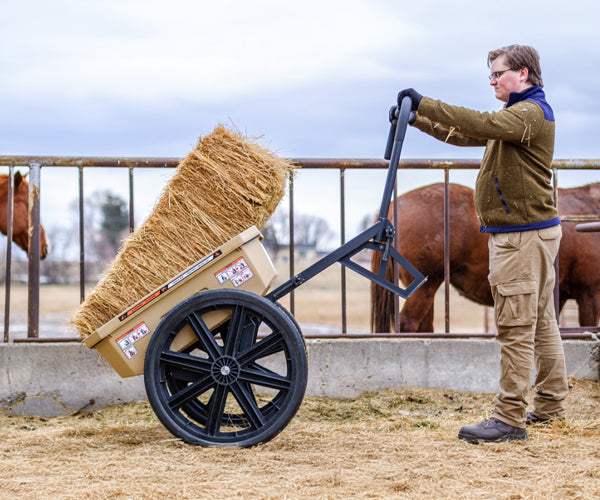 A farm worker tips the cart up to unload a hay bale from the cart as horses stand in the background.