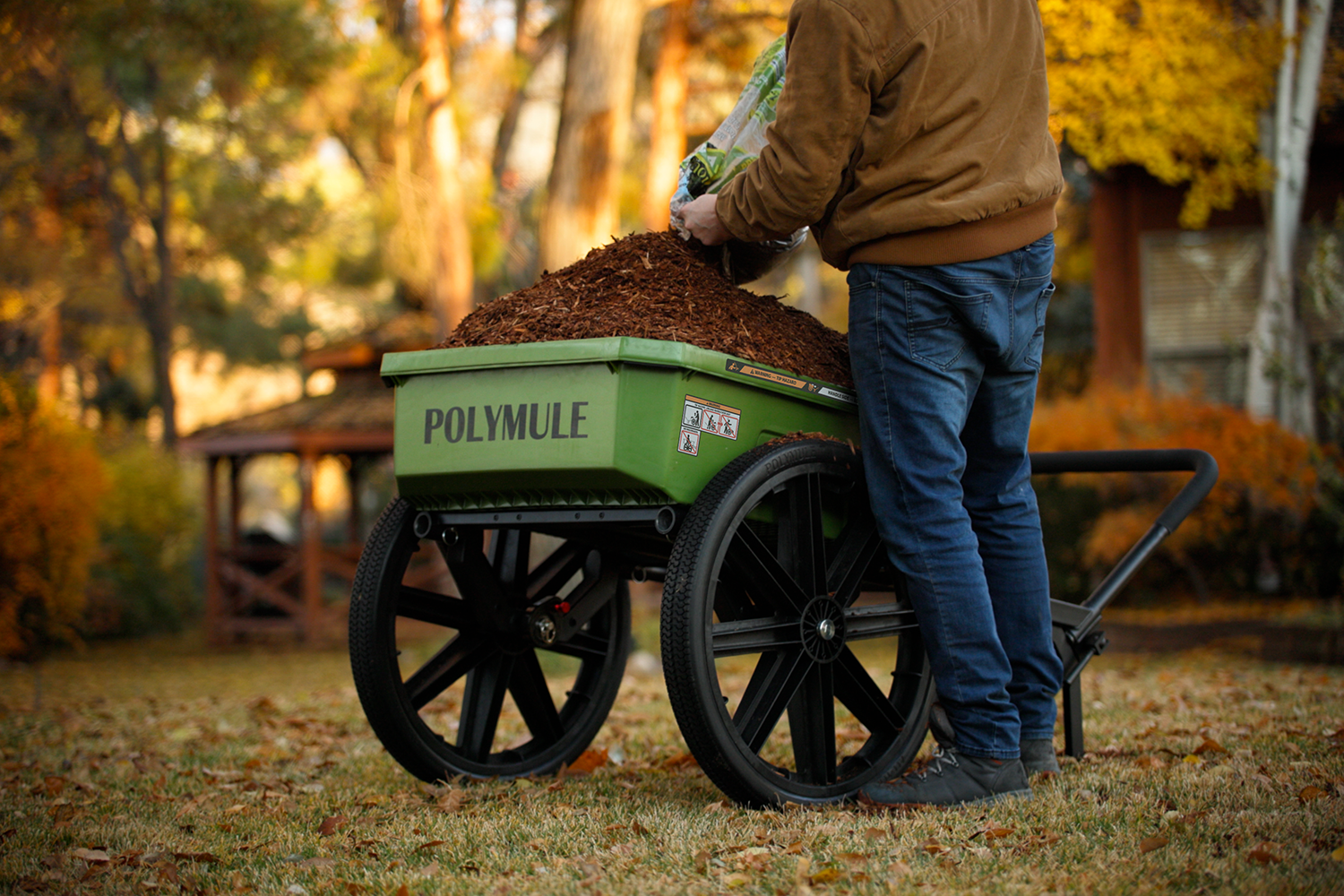 A man fills a green Warrior cart with mulch from a bag in front of a cabin in the woods. 